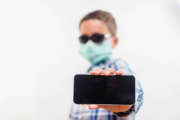 Young boy wearing disposable mask and sunglasses holding mobile phone over white background - Φωτογραφία, εικόνα