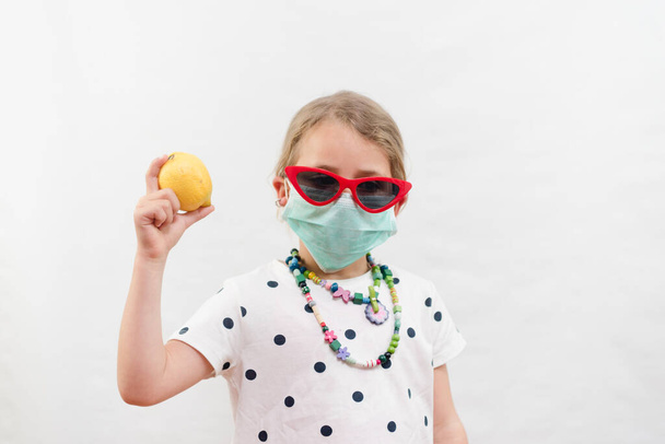 Little blond girl wearing disposable mask and sunglasses holding yellow lemon vitamin C source over white background - Foto, Bild