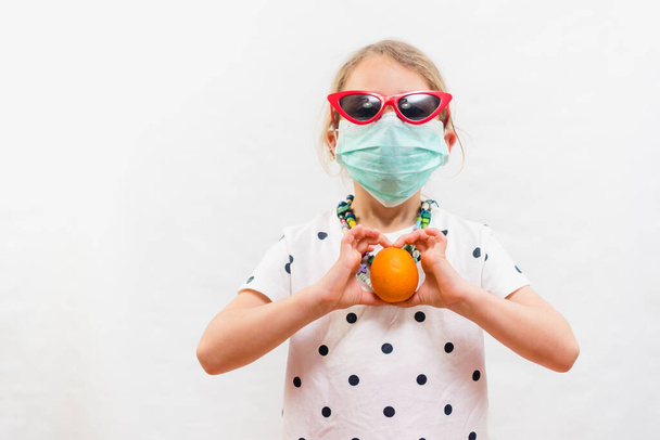 Little blond girl wearing disposable mask and sunglasses holding an orange vitamin C source over white background - Fotoğraf, Görsel