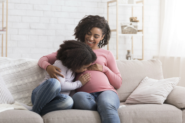 Schattig klein Afrikaans meisje zoenen en knuffelen haar zwangere moeders buik - Foto, afbeelding