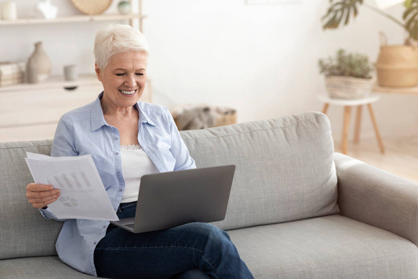glimlachende oudere vrouw werken met laptop en documentatie thuis - Foto, afbeelding
