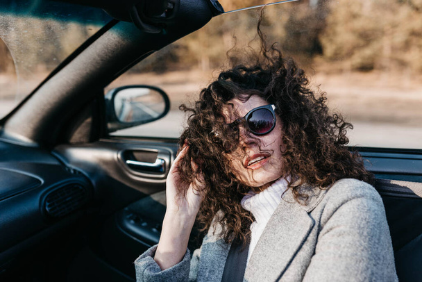 Curly woman photographer in a convertible car rides on a sunny day on the road and rejoices in sunglasses - Fotó, kép