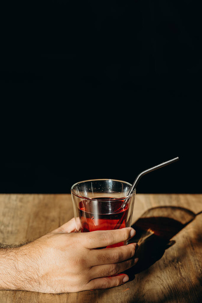 Una mano de hombre sosteniendo un vaso de bebida dulce roja con paja de metal o paja inoxidable en la mesa de madera
. - Foto, imagen