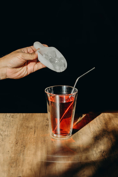 金属製の藁や木のテーブルの上にステンレス製のわらで赤い甘い飲み物のガラスの上に測定スプーン付きのアイスキューブを保持男の手. - 写真・画像