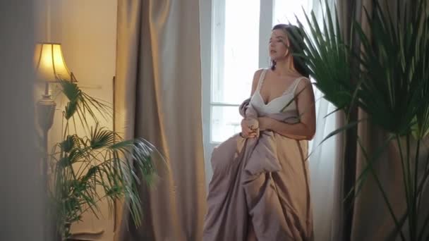 Beautiful young caucasian brunette bride in luxurious white lingerie covered with blanket walks around hotel bedroom and enjoys morning on the background of indoor palm flowers, luminous floor lamp. - Imágenes, Vídeo