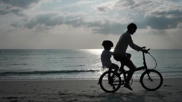 Aziatische familie Op zomervakantie Vader en zoon fietsen op het strand Vakantie en Reizen concept. Silhouet Slow Motion - Video
