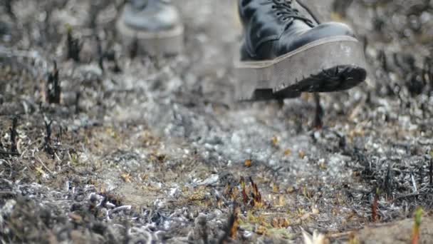 Fille moderne en cuir à la mode bottes noires avec semelles hautes marche le long de la cendre brûlée de l'herbe dans un champ inhabité et s'arrête, se concentrant sur ses pieds avec des chaussures, vue du bas au ralenti
. - Séquence, vidéo
