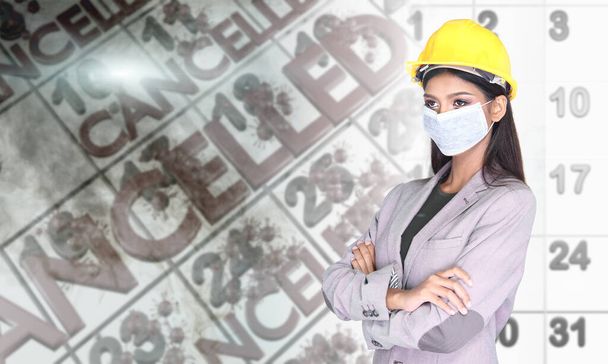 Engineer Woman in Gray Suit wear Yellow Safety Hard Hat and industrial surgical dust mask, portrait over background of 3d render coronavirus or covid-19 as job cancellation on work schedule calendar - Photo, Image