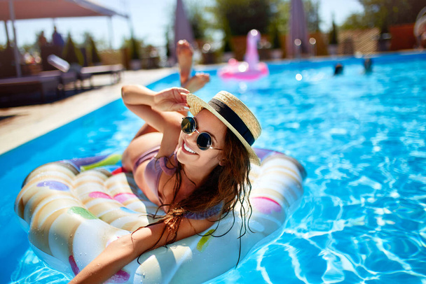 Young smiling fitted girl in bikini, straw hat relax on inflatable swan in swimming pool. Attractive woman in swimwear lies in the sun on tropical vacation. Pretty female sunbathing at luxury resort. - Foto, immagini