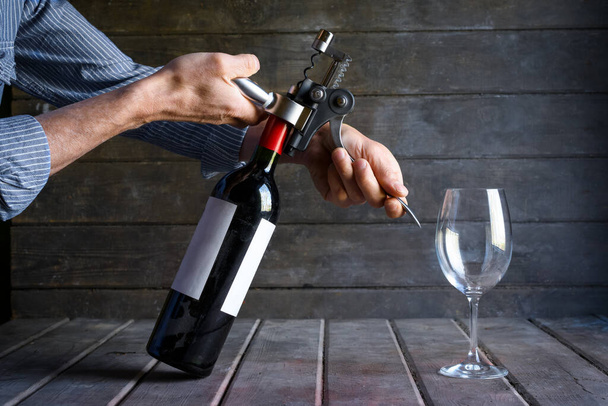 Manos masculinas abriendo una botella de vino con un sacacorchos. Una botella polvorienta de vino tinto y una copa sobre el telón de fondo de viejas tablas de madera
. - Foto, Imagen