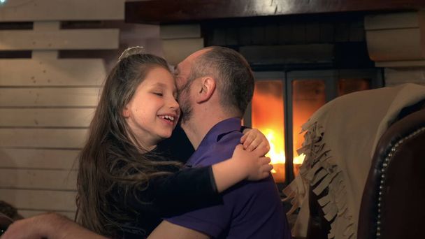 Счастливый отец и маленькая дочь обнимают друг друга на кресле возле камина
 - Фото, изображение