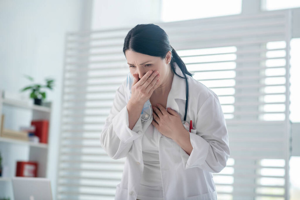 Woman in white coat and stethoscope standing coughing - Fotoğraf, Görsel