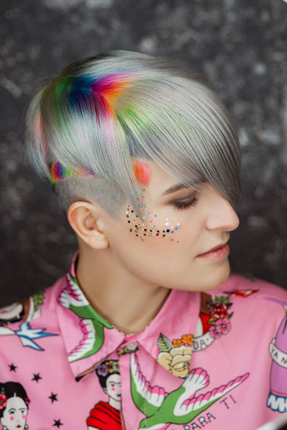 Portrait of a young beautiful girl in a pink shirt on a gray background with dyed hair. Short haircut pixie or bob. Rainbow coloring and sequins on the face. - Фото, зображення