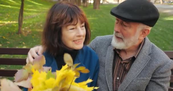 Cheerful couple of senior citizens man and woman are sitting relaxing on a bench in a cozy park in autumn. Bearded handsome elderly husband jokes, hugs and kisses his wife with a bouquet of leaves. - Footage, Video