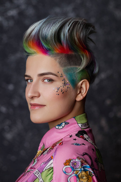 Portrait of a young beautiful girl in a pink shirt on a gray background with dyed hair. Short haircut pixie or bob. Rainbow coloring and sequins on the face. - Foto, imagen