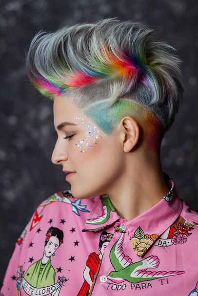 Portrait of a young beautiful girl in a pink shirt on a gray background with dyed hair. Short haircut pixie or bob. Rainbow coloring and sequins on the face. - 写真・画像