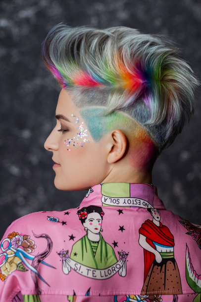 Portrait of a young beautiful girl in a pink shirt on a gray background with dyed hair. Short haircut pixie or bob. Rainbow coloring and sequins on the face. - Fotó, kép