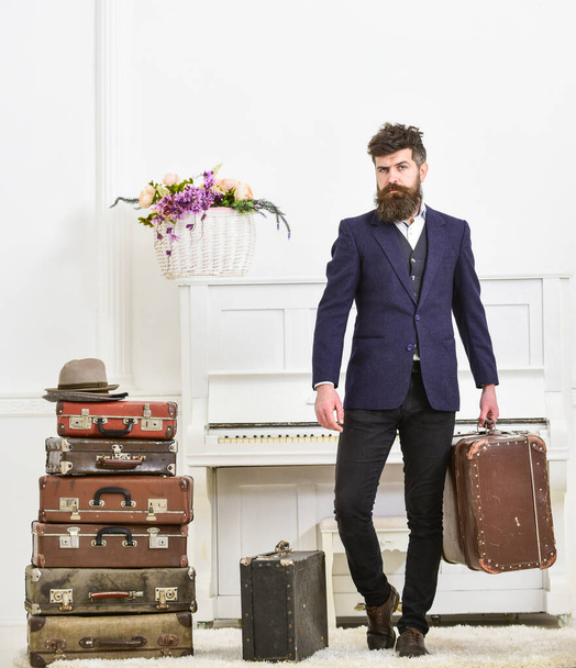 Macho attractive, elegant on strict face carries vintage suitcases. Butler and service concept. Man with beard and mustache wearing classic suit delivers luggage, luxury white interior background. - Fotoğraf, Görsel