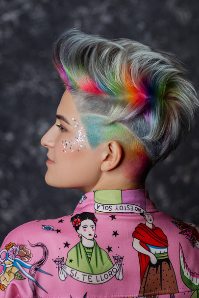 Portrait of a young beautiful girl in a pink shirt on a gray background with dyed hair. Short haircut pixie or bob. Rainbow coloring and sequins on the face. - Photo, Image