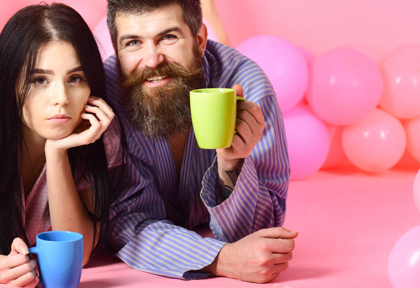 Man and woman on smiling faces lay, pink background. Couple relax in morning with coffee. Weekend morning concept. Couple in love drink coffee in bed. Man and woman in domestic clothes, pajamas. - Photo, image