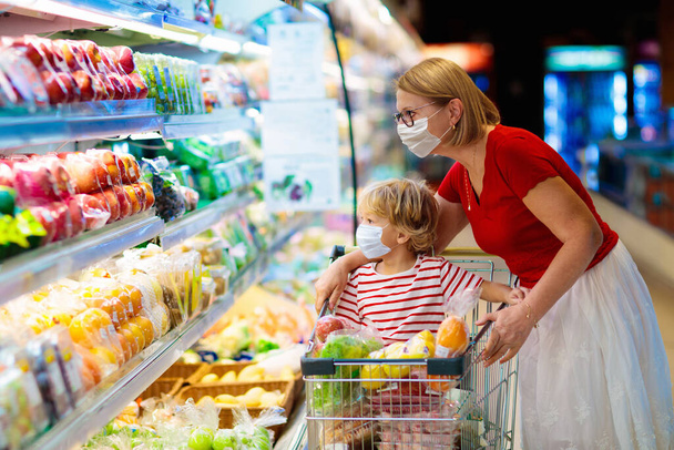 Zakupy z dziećmi podczas epidemii wirusa. Matka i dziecko w masce chirurgicznej kupują owoce w supermarkecie. Mama i mały chłopiec kupują świeże warzywa w spożywczaku. Rodzina w sklepie.  - Zdjęcie, obraz
