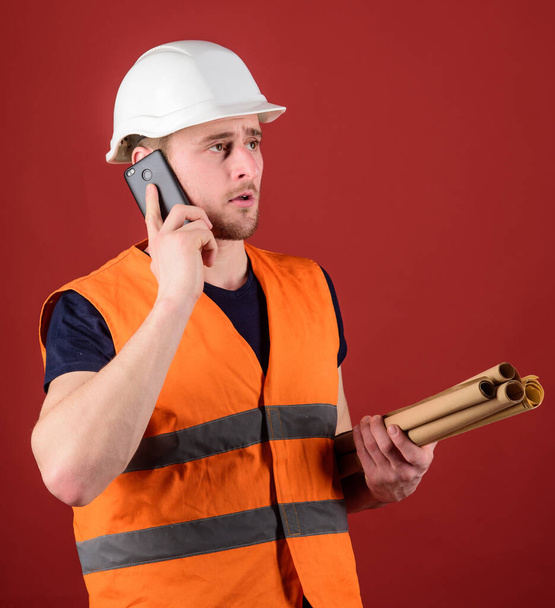 Architect concept. Engineer, architect, builder on busy face speaks on smartphone while holds blueprints. Man, foreman in helmet supervises construction on phone, red background. - Photo, image