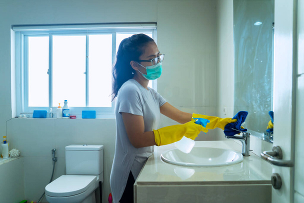 Portrait of young female cleaning the bathroom using disinfectant while wearing gloves - 写真・画像