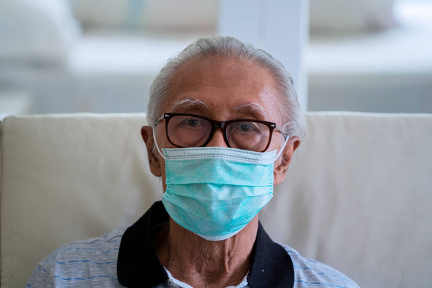 Portrait of grandfather looking at camera while wearing facial mask and sitting on sofa  - Valokuva, kuva