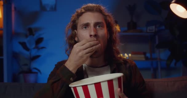 Close up view of handsome man with long hair watching sport and eating popcorn. Millennial hipster guy sitting on sofa and looking at tv screen with unsatisfied face. Concept of leisure. - Video