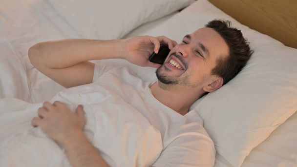 Happy Young Man Talking on Smartphone in Bed - 写真・画像