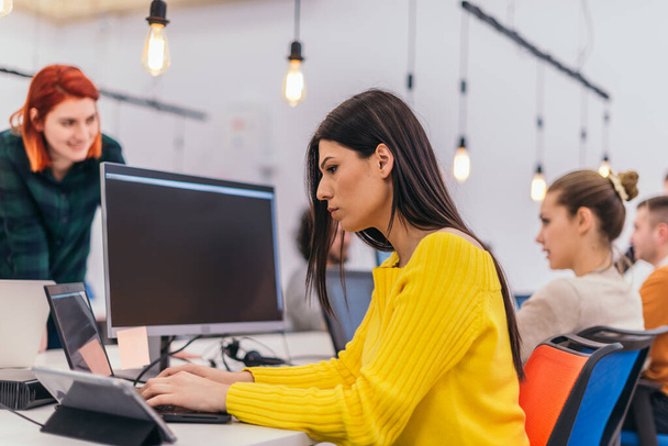 Konzentrierte schöne Mädchen mit langen schwarzen Haaren und gelber Bluse bei der Arbeit an einem Laptop in einem modernen Büro. - Foto, Bild