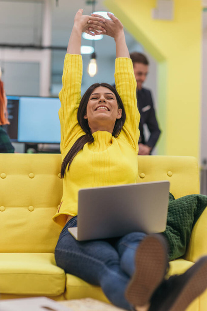 Schöne Geschäftsfrau (Angestellte) sitzt auf einer gelben Couch, während sie an ihrem Laptop in einem Start-up-Unternehmen arbeitet. - Foto, Bild