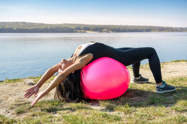 Mujer joven en forma en ropa deportiva durante el tiempo de fitness y hacer ejercicio con la pelota en el lago. Estilo de vida saludable - Foto, imagen