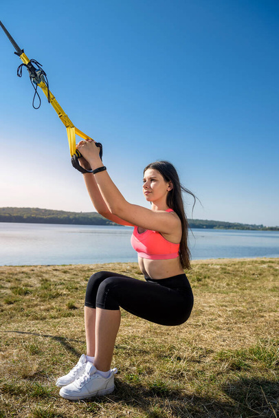 Attractive young  woman  doing TRX training outdoors near the lake at daytime. Healthy lifestyle - 写真・画像