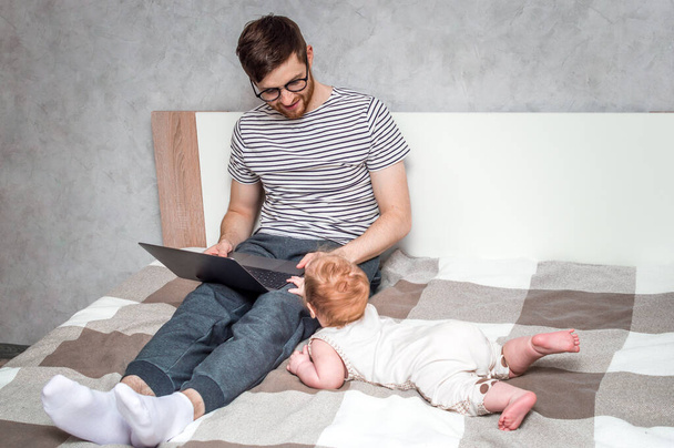 Young man working on a laptop in bed at home and baby beside. Concept Working remotely - Foto, Imagem