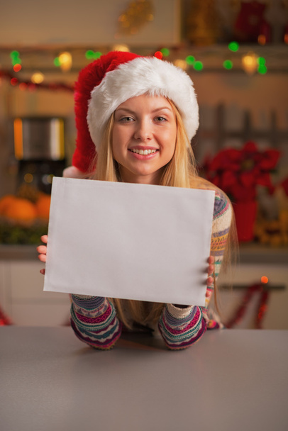 Teenage girl in santa hat showing blank paper sheet in chrristmas decorated kitchen - Foto, imagen