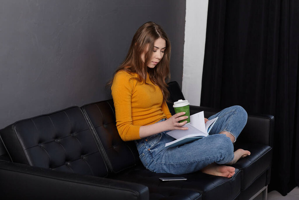 Ein junges Mädchen sitzt zu Hause auf einem schwarzen Ledersofa, hält eine Tasse Kaffee in der Hand und liest Notizen in einem Notizbuch. Arbeit zu Hause - Foto, Bild