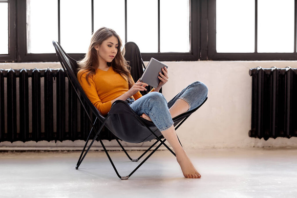Młoda dziewczyna siedzi w domu na stylowym czarnym skórzanym krześle, trzyma filiżankę kawy i tablet w ręku. Praca i relaks w domu - Zdjęcie, obraz