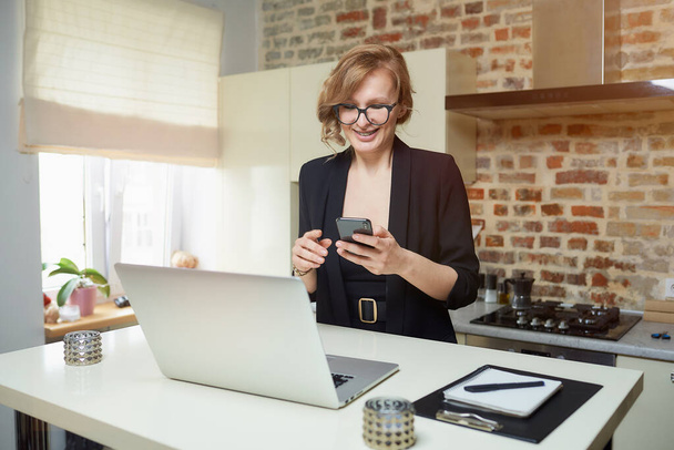 A blond woman in glasses works remotely in her kitchen. A happy girl with braces browses news on the internet with her smartphone at home. A teacher with a smile texting to her remote students. - Photo, Image