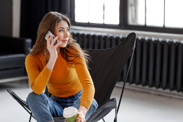 Ένα νεαρό κορίτσι κάθεται σε μια κομψή μαύρη δερμάτινη καρέκλα, κρατά ένα φλιτζάνι καφέ και ένα τηλέφωνο στο χέρι της. Εργασία, επικοινωνία, χαλάρωση στο σπίτι - Φωτογραφία, εικόνα