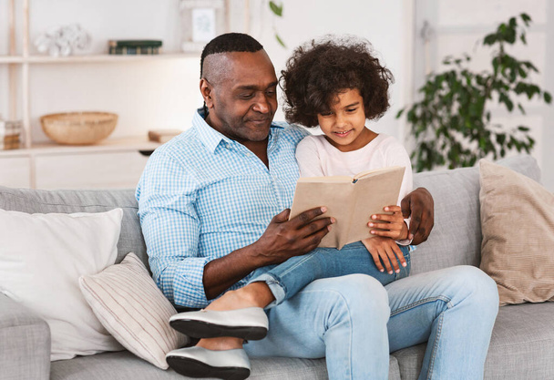 Interesujące spędzanie czasu w domu. Pretty African American dziewczyna słuchając dziadka czytać książkę w salonie - Zdjęcie, obraz