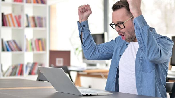 Young Man in Glasses Celebrating Success on Laptop - 写真・画像
