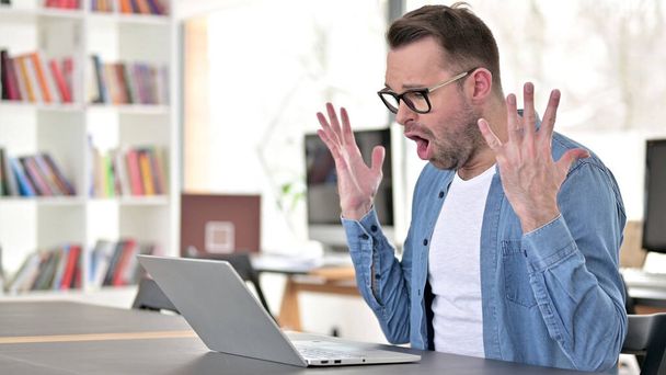 Młody człowiek w okularach zdenerwowany utratą, przy użyciu laptopa - Zdjęcie, obraz