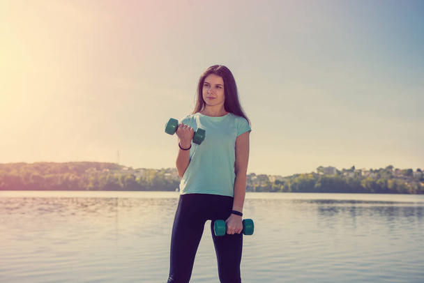 young beautiful girl is engaged in morning gymnastics with dumbbells at the lake - Photo, Image