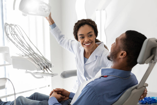 Mooie zwarte dame tandarts groeten mannelijke patiënt op haar werkplek - Foto, afbeelding