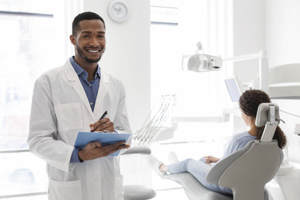 Czarny dentysta uśmiechający się i trzymający folder w gabinecie stomatologicznym - Zdjęcie, obraz