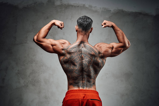Maskuliner Sportler isoliert auf dem grauen Hintergrund in Sporthose, zeigt seine Rückentätowierung und Muskeln, während er die Arme in die Höhe reckt - Foto, Bild