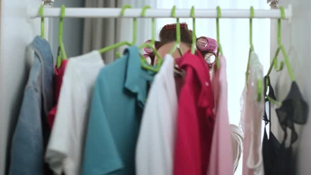 Beautiful young girl in morning in curlers on her head and in housecoat opens wardrobe, selects things dresses and t-shirts for trip to street, does not choose anything closes dressing room. - Кадры, видео