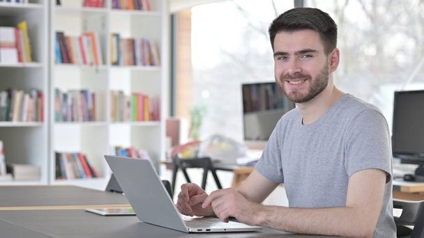 Dizüstü bilgisayarlı genç adam ofiste kameraya gülümsüyor. - Fotoğraf, Görsel