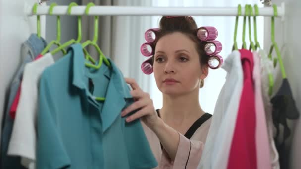 Beautiful young girl in morning in curlers on her head and in housecoat opens wardrobe and chooses blue evening dress for going out for walk. Woman measures dress near mirror and closed dressing room. - Séquence, vidéo